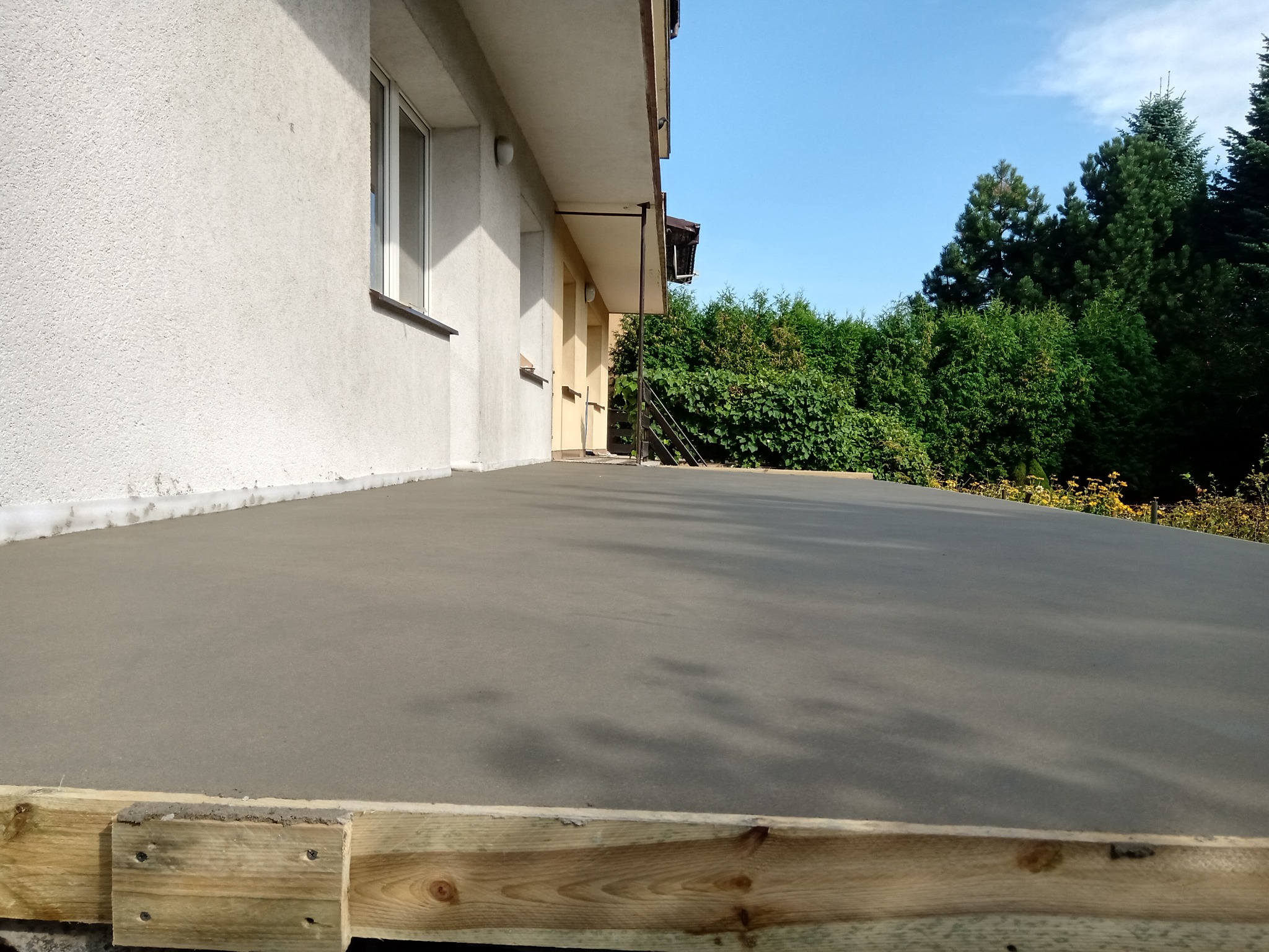 Świeżo wylana, gładka betonowa wylewka na balkonie z widocznym drewnianym szalunkiem, na tle ściany budynku i zieleni.