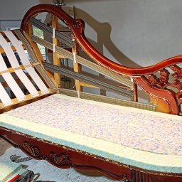 Marek Krajnik - Rozłożona sofa w stylu retro z drewnianą ramą, widoczne sprężyny, pianka i pasy. Etap prac renowacyjnych, narzędzia w tle. Ciemne drewno, jasne materiały.