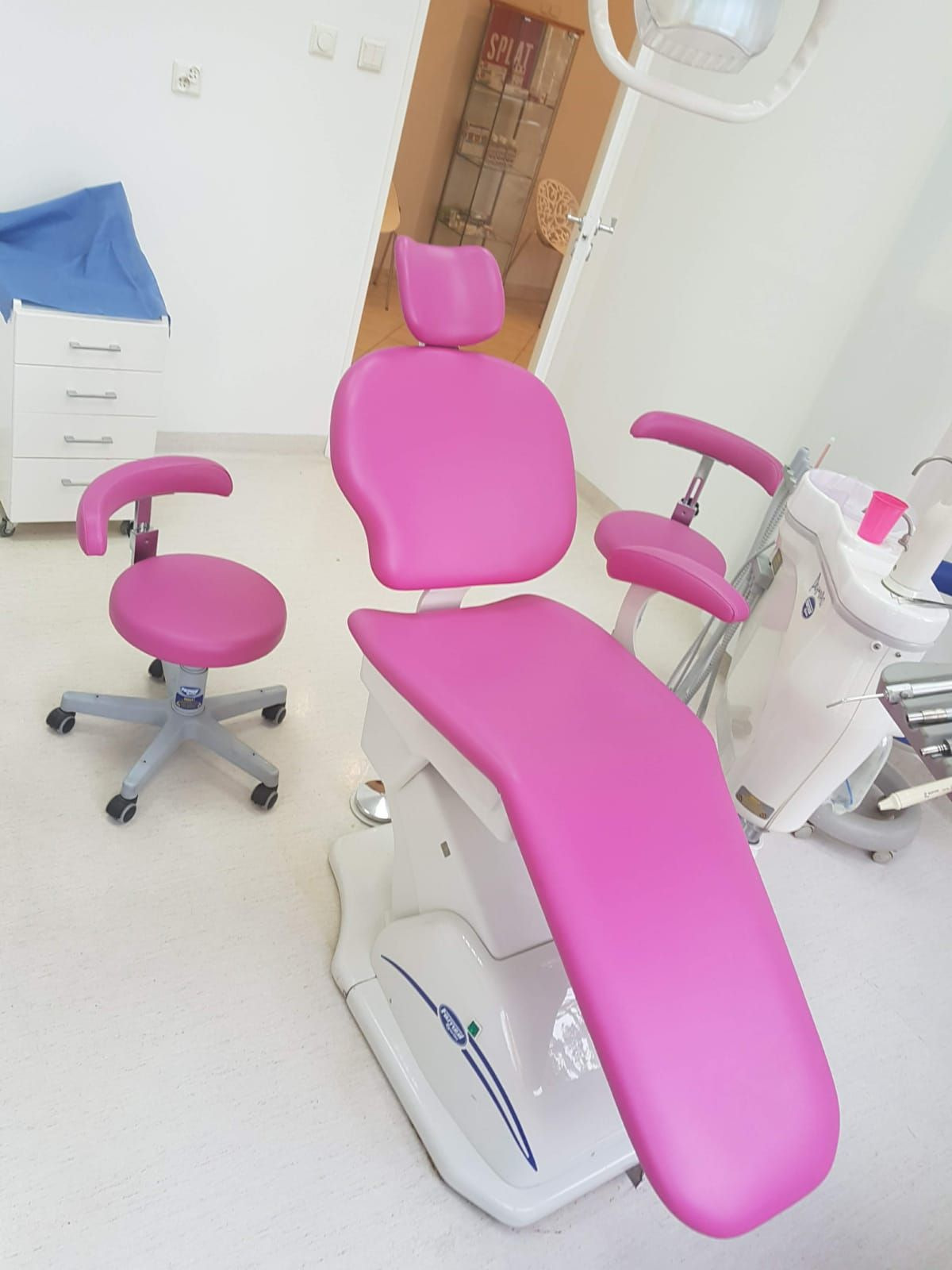 Różowe obicie fotela dentystycznego z krzesłem na kółkach w jasnym gabinecie. Widoczne białe szafki i sprzęt dentystyczny. Nowoczesny design, czyste wnętrze.