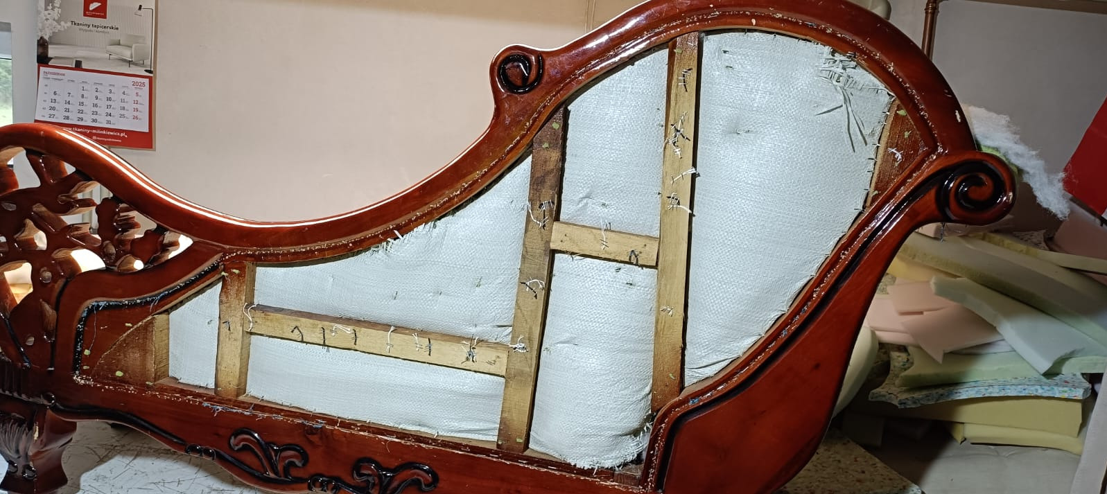 Szkielet sofy w trakcie renowacji: drewniana konstrukcja, zszyta tkanina, widoczne zszywki. Elementy dekoracyjne z ciemnego drewna. W tle kalendarz.