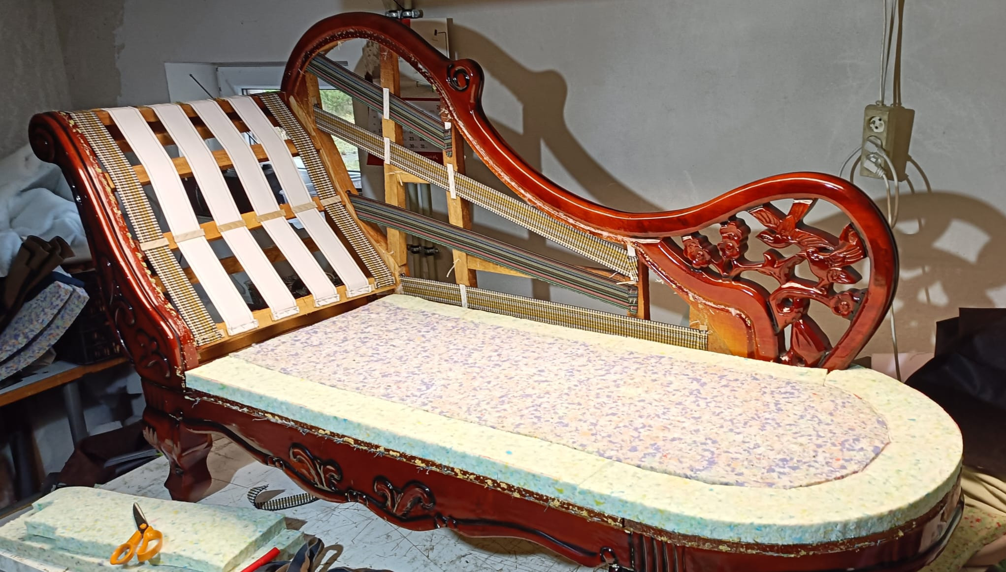 Rozłożona sofa w stylu retro z drewnianą ramą, widoczne sprężyny, pianka i pasy. Etap prac renowacyjnych, narzędzia w tle. Ciemne drewno, jasne materiały.