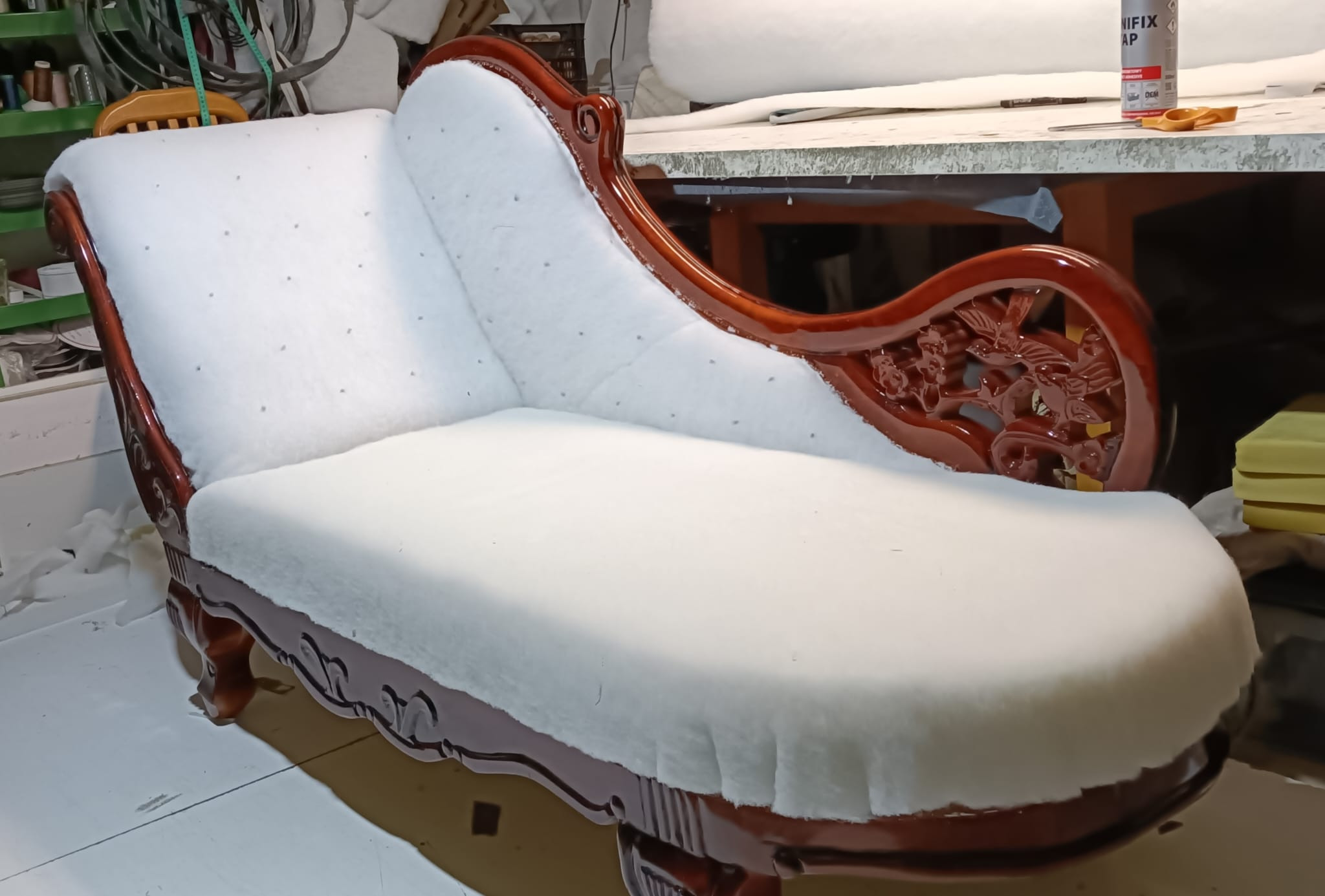 Antyczna leżanka z rzeźbionym, lakierowanym drewnem w trakcie obijania białą watoliną. Widoczne narzędzia i materiały tapicerskie w tle. Detal rzemiosła.