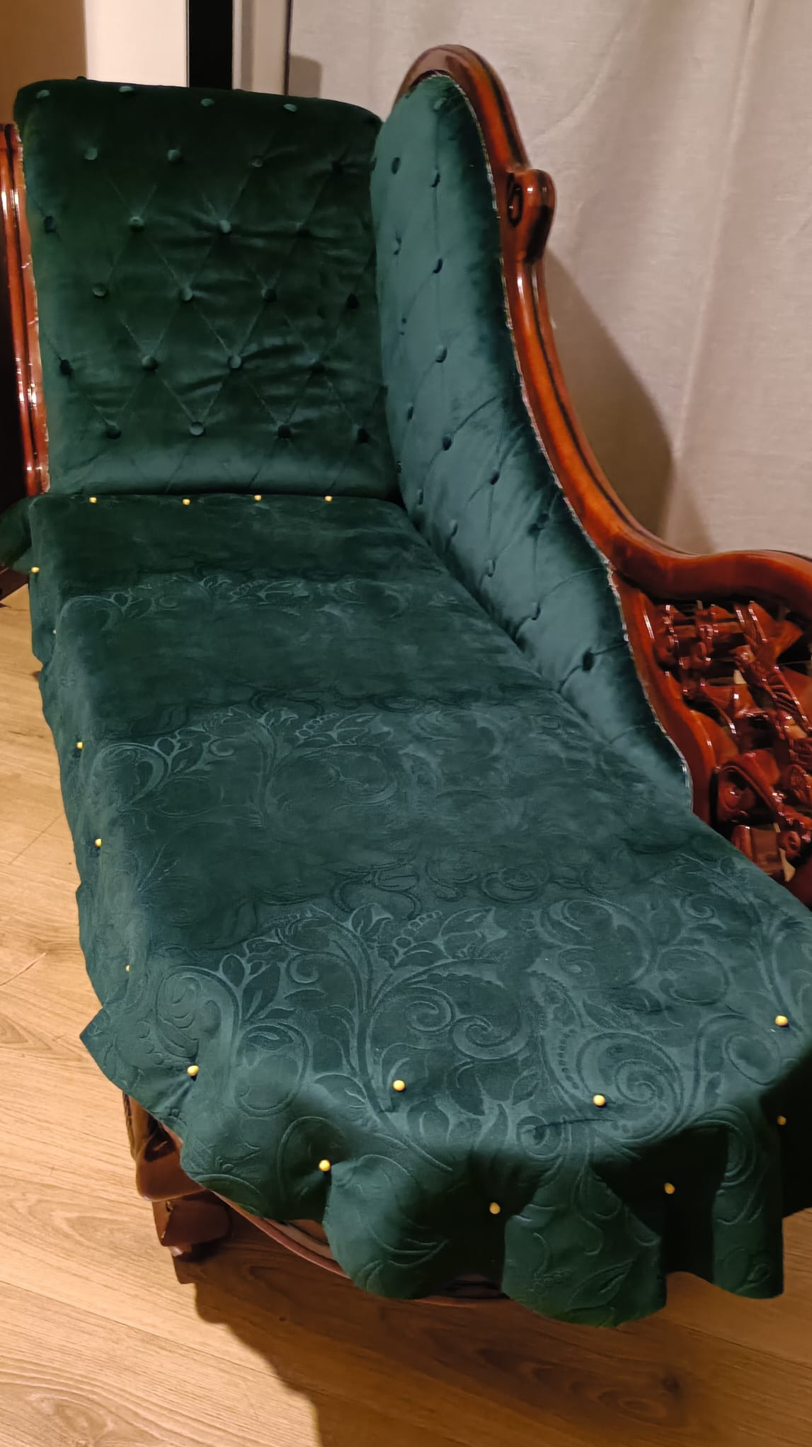 Szmaragdowy szezlong z pikowanym oparciem i drewnianymi zdobieniami, w trakcie prac z użyciem szpilek, ukazujący etap dopasowywania tkaniny.