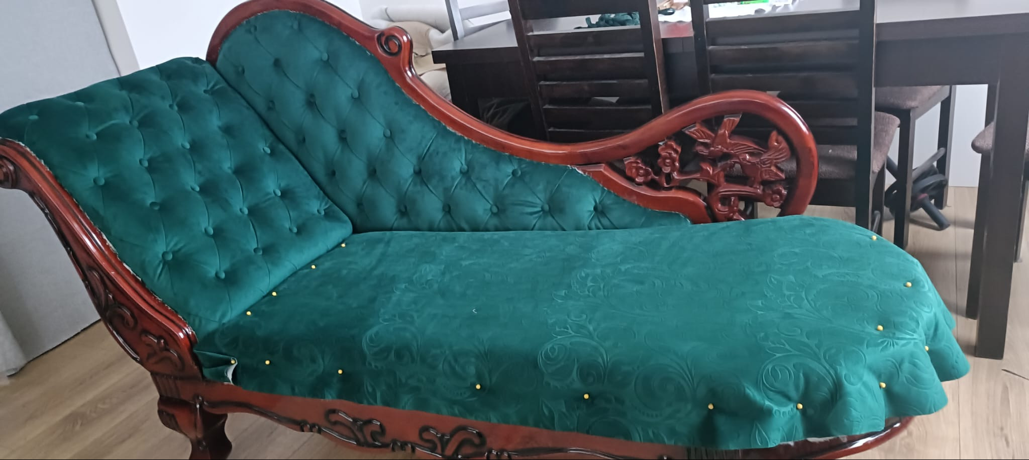 Stylowy szezlong z ciemnozieloną, wzorzystą tapicerką i drewnianą ramą z rzeźbieniami. Widoczne złote pinezki mocujące materiał. W tle stół i krzesła.