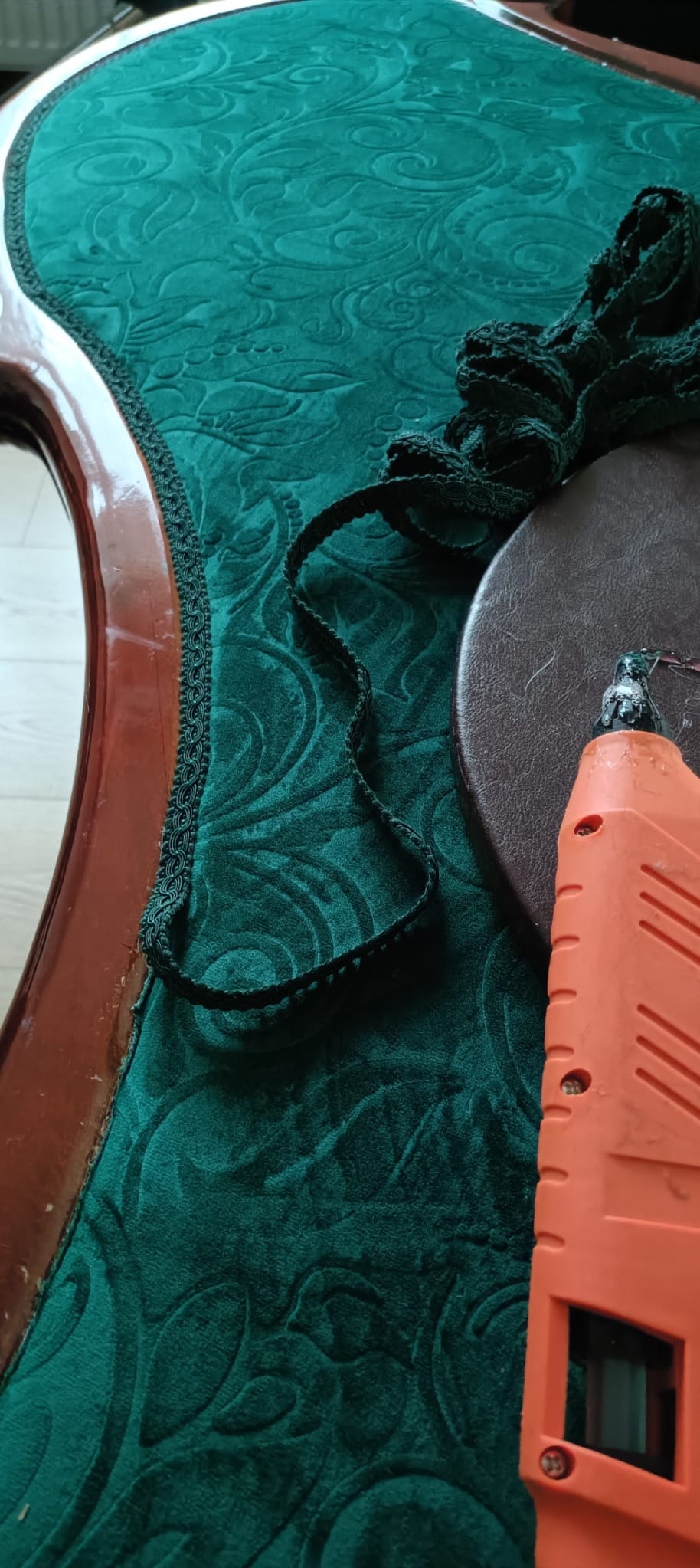 Zielone obicie krzesła z wzorem, czarna ozdobna taśma, siedzisko i pistolet do kleju. Praca w toku, detale wykończenia, narzędzia pracy.