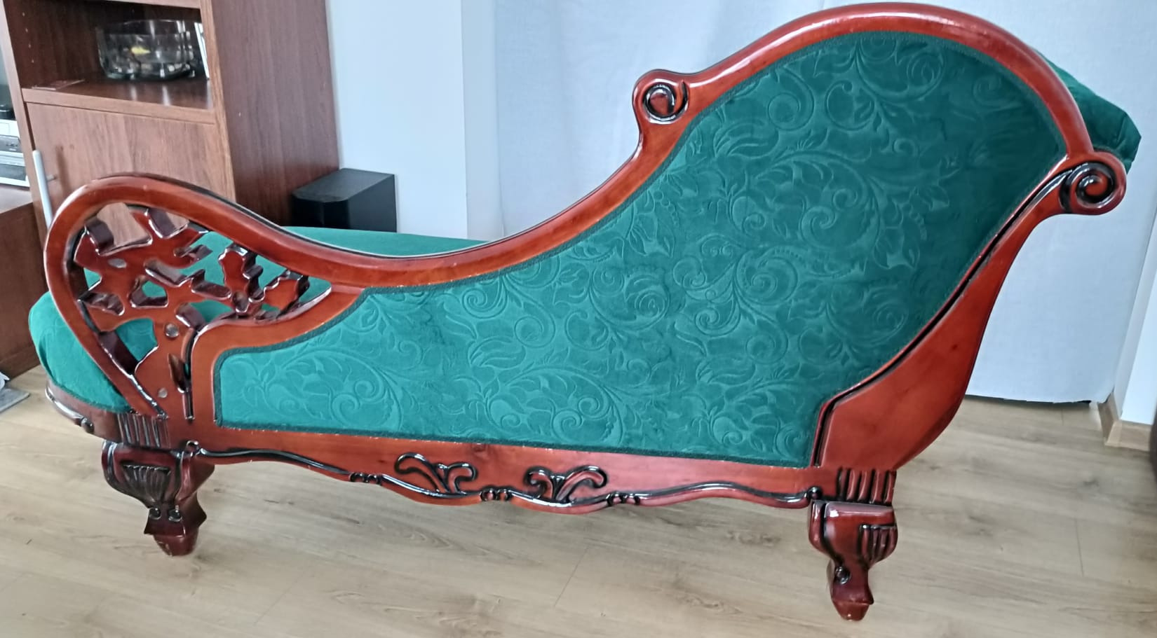 Stylowy szezlong z drewnianą ramą i zieloną, wzorzystą tapicerką. Widoczne ozdobne nogi i wykończenia. Klasyczny design.