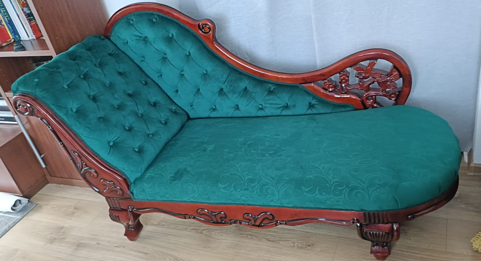 Stylowy szezlong z ciemnozieloną, aksamitną tapicerką i rzeźbioną, drewnianą ramą w kolorze wiśni. Widoczne pikowania i dekoracyjne wykończenia.