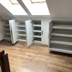 Biała zabudowa meblowa pod skosem na poddaszu z oknami dachowymi, widoczne otwarte szafki z półkami i szufladami, poziomica na krześle w rogu kadru.
