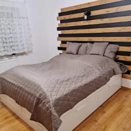 Sypialnia z ł&oacute;żkiem na białej ramie z szufladami, przykrytym brązową narzutą i poduszkami, z drewniano-czarnym zagł&oacute;wkiem w poziome pasy oraz lampką na ścianie.