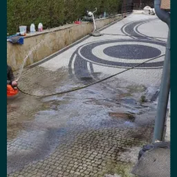 Czyszczenie kostki brukowej z wzorem w kształcie okręgów za pomocą węża ciśnieniowego; widoczne mokre powierzchnie i osoba w pomarańczowym ubraniu.