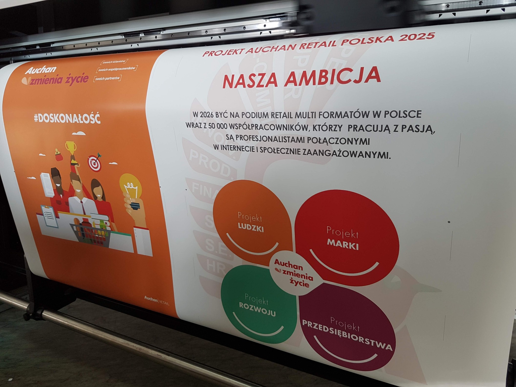 Drukarka wielkoformatowa w trakcie drukowania plakatu z hasłem 'Nasza ambicja' i elementami identyfikacji wizualnej Auchan Retail Polska 2025, widoczne kolorowe grafiki i tekst.
