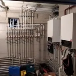 Dwa białe piece centralnego ogrzewania zamontowane na ścianie w pomieszczeniu z licznymi rurami instalacyjnymi i zaworami, widoczne narzędzia i pojemnik, instalacja wykonana w Rumi.