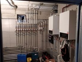 Dwa białe piece centralnego ogrzewania zamontowane na ścianie w pomieszczeniu z licznymi rurami instalacyjnymi i zaworami, widoczne narzędzia i pojemnik, instalacja wykonana w Rumi.