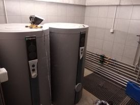Dwa szare, cylindryczne zbiorniki akumulacyjne Viessmann w kotłowni z jasnymi, kafelkowymi ścianami. Widoczne rury i elementy instalacji. Na jednym ze zbiorników leży poziomica.