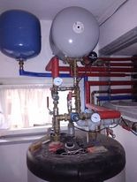Złożona instalacja grzewcza z dwoma zbiornikami (niebieski i szary), rurami miedzianymi, zaworami, manometrami oraz pompą obiegową, widoczna na tle białej ściany i okna.