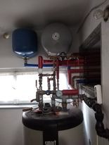 Kocioł grzewczy z podłączonymi rurami instalacji centralnego ogrzewania, zbiornik wyrównawczy w kolorze niebieskim i szarym, widoczne manometry i zawory, fragment okna w tle.