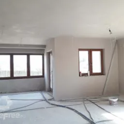 Pokój po nałożeniu gładzi maszynowej na ściany i sufit, widoczne wystające kable elektryczne, okna z brązowymi ramami, wiadro z resztkami gładzi na podłodze.