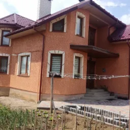 Dwupiętrowy dom z pomarańczową elewacją o fakturze baranka, brązową stolarką okienną i bordową dachówką, widok z poziomu ogródka warzywnego w słoneczny dzień.