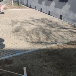 Ułożona kostka brukowa na posesji, widoczne cienie drzew na nawierzchni, obok przygotowany teren pod trawnik i leżąca szczotka do zamiatania.