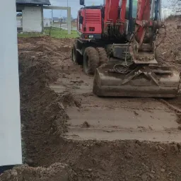 Czerwona koparka kołowa wykonuje wykop pod fundamenty obok białego budynku w pochmurny dzień.