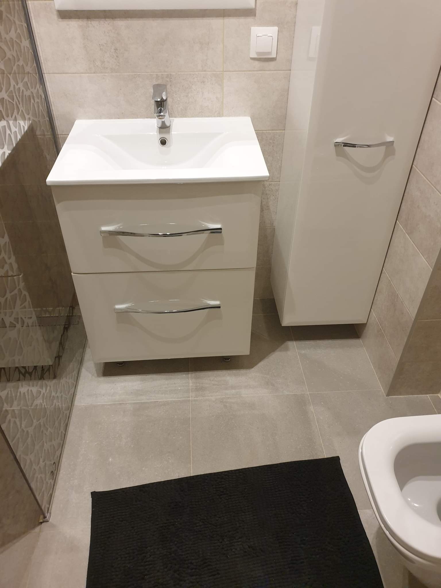 Nowoczesna, jasna łazienka z białą szafką umywalkową z dwiema szufladami z chromowanymi uchwytami, wysoką białą szafką wiszącą, szarymi płytkami i czarnym dywanikiem.