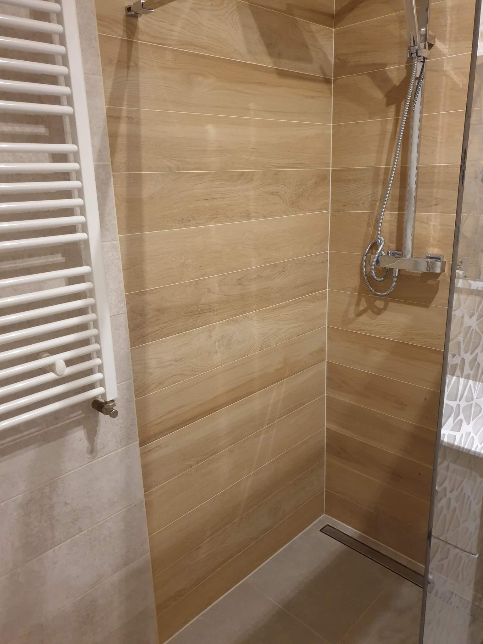 Kabina prysznicowa wyłożona płytkami imitującymi drewno, z odpływem liniowym w podłodze i zamontowaną baterią prysznicową z deszczownicą, obok grzejnik drabinkowy.
