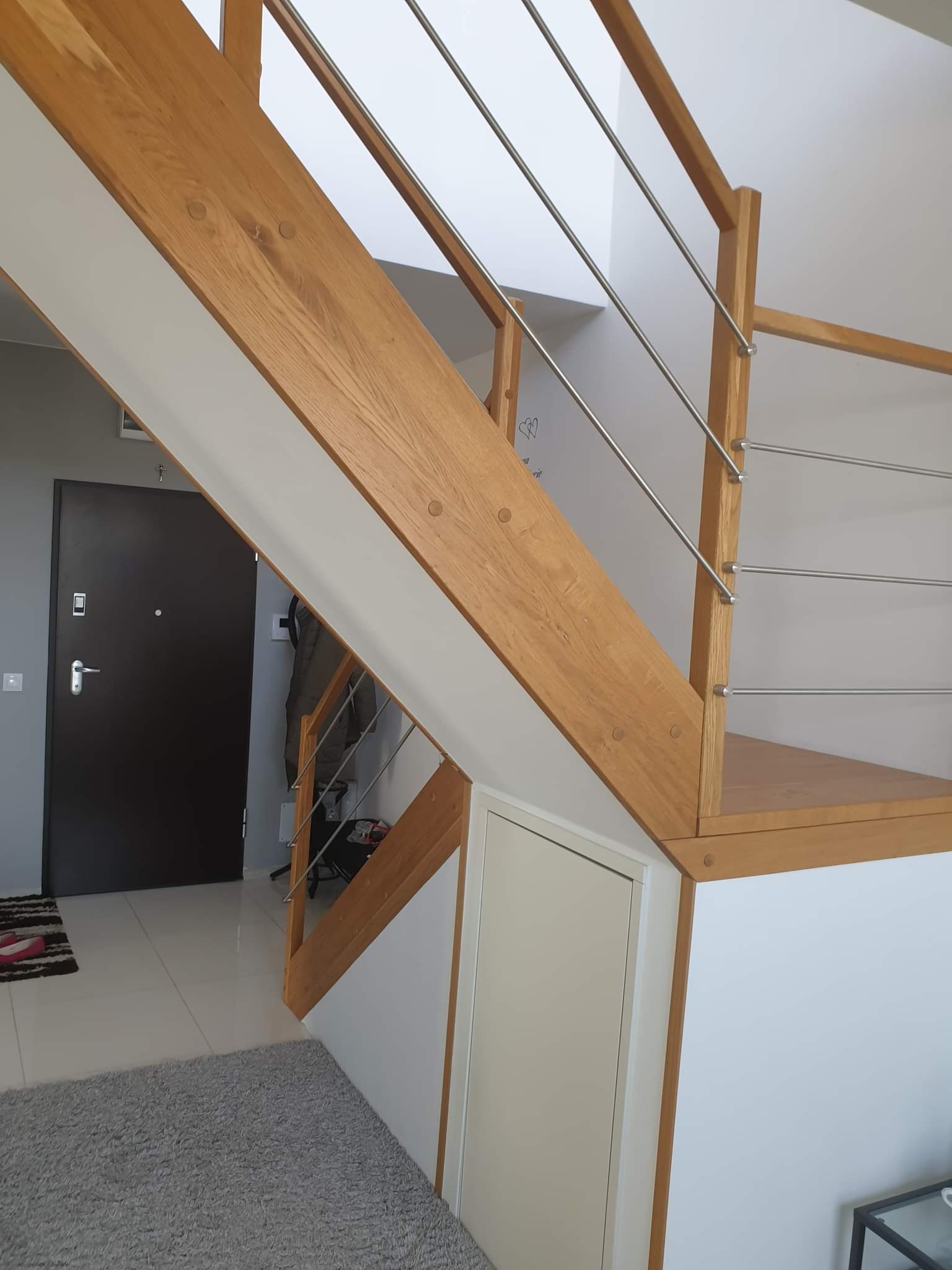 Drewniane schody z metalową balustradą w nowoczesnym wnętrzu, widoczne wejście do pomieszczenia z ciemnymi drzwiami, jasna podłoga i szary dywan.