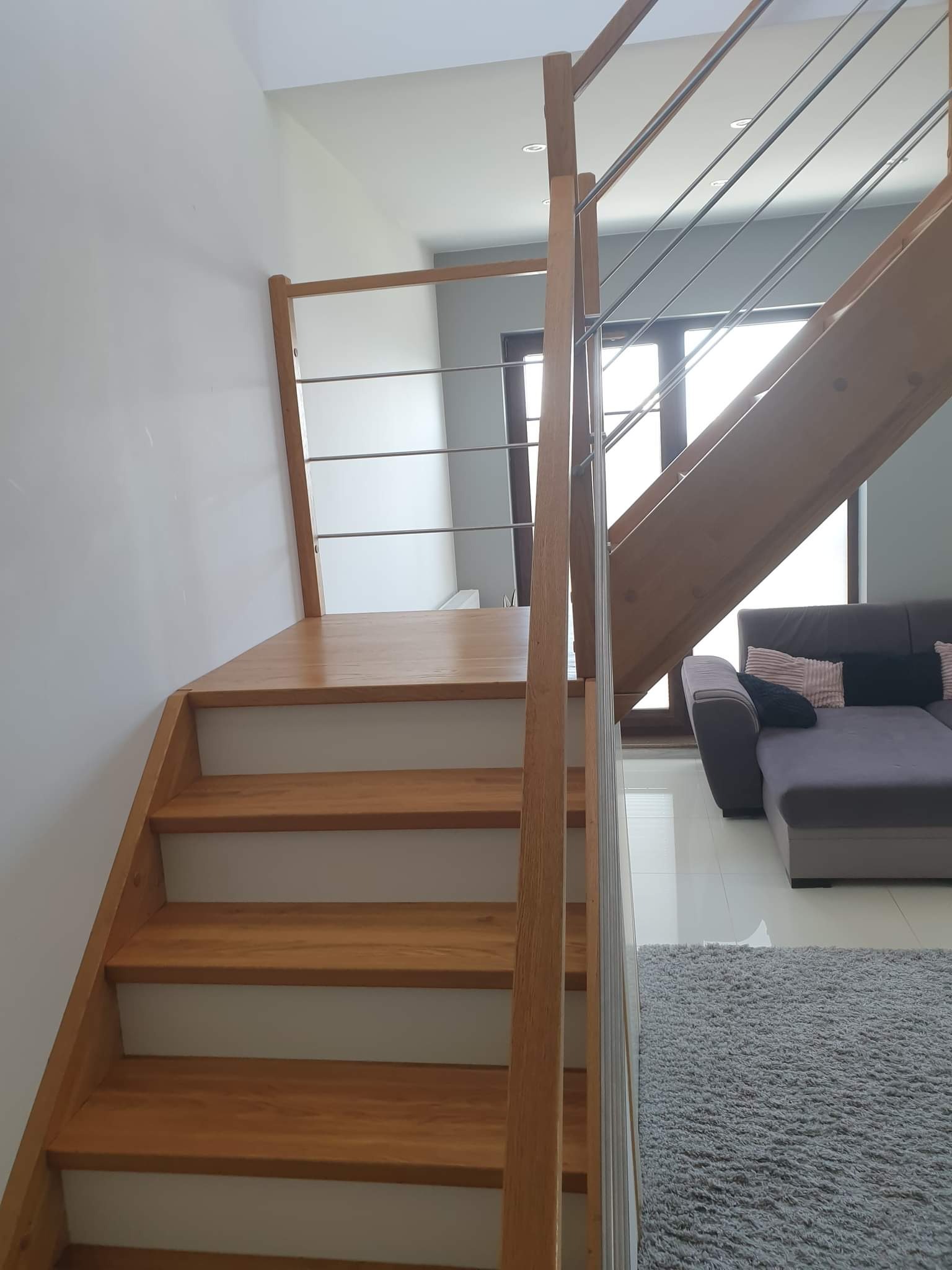 Drewniane schody z metalową balustradą w nowoczesnym wnętrzu, widok z dołu, szare ściany i dywan, sofa w tle.