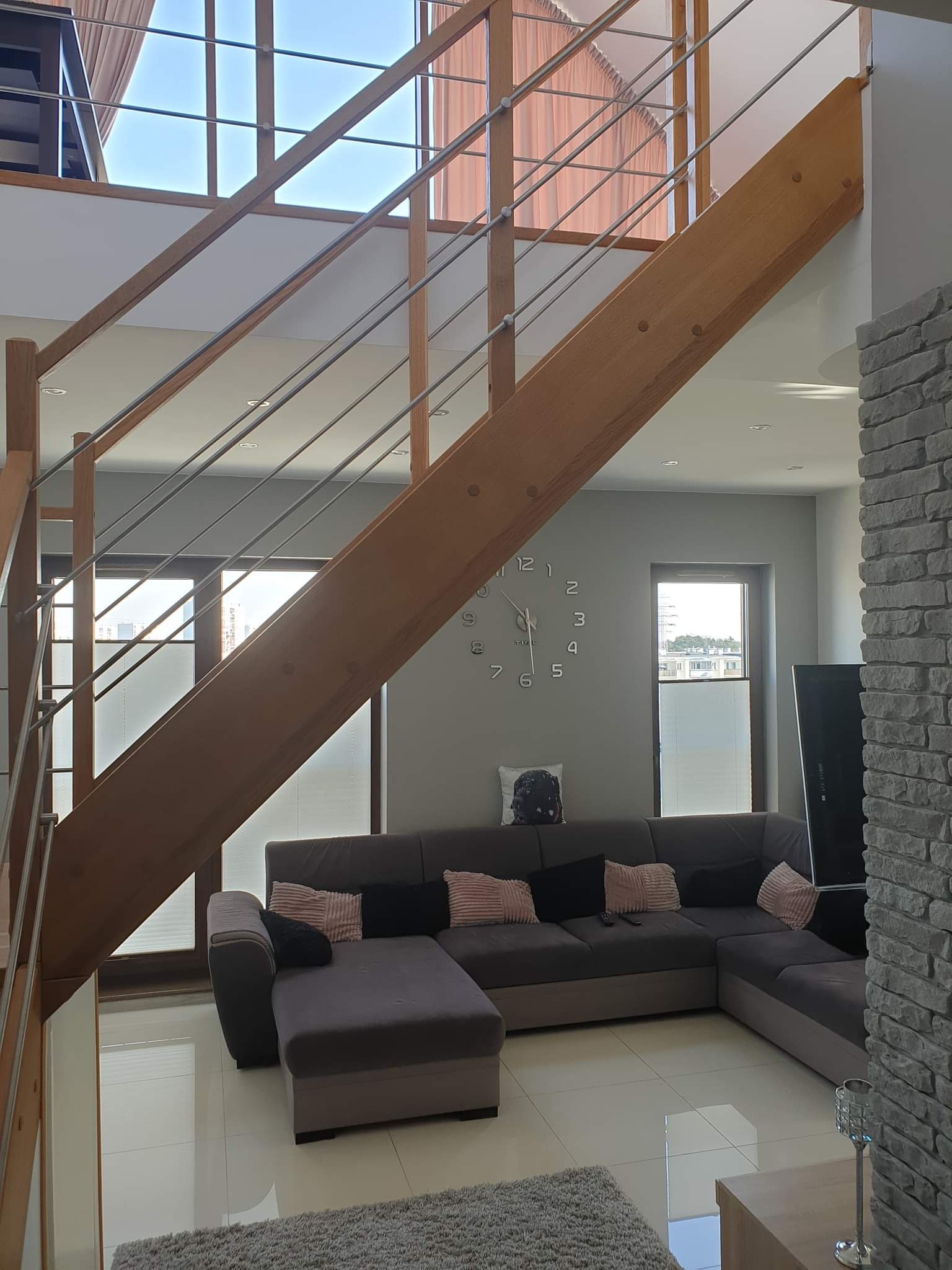 Dwupoziomowy salon z drewnianymi schodami z metalowymi barierkami, szarą sofą narożną z poduszkami, zegarem ściennym i ścianą z dekoracyjnej cegły.