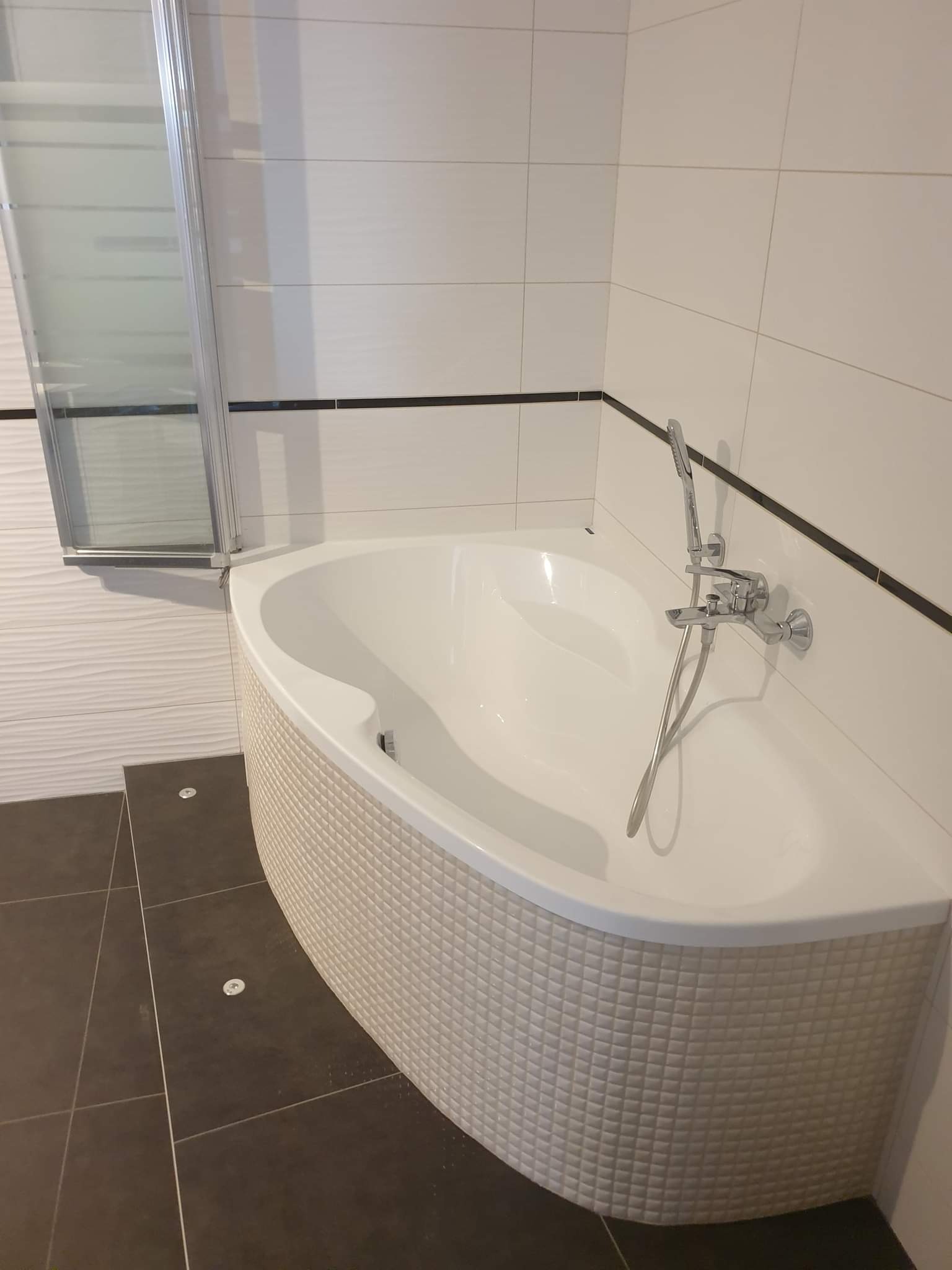 Narożna wanna z obudową w mozaikę, chromowana bateria z prysznicem, szklany parawan nawannowy, ciemna podłoga z płytkami i punktowe oświetlenie, białe płytki na ścianach z czarnym akcentem.