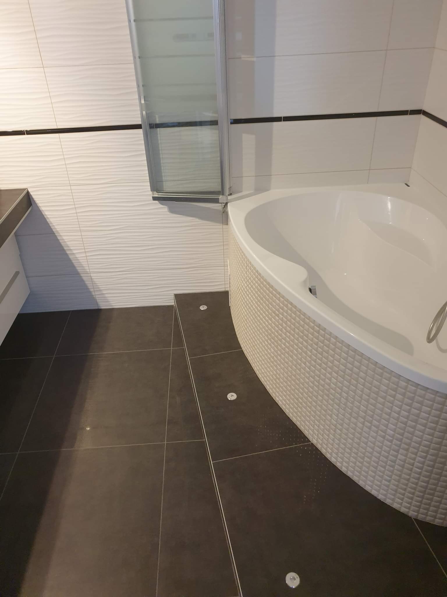 Nowoczesna łazienka w Krakowie z narożną wanną obudowaną mozaiką, grafitową podłogą z płytek i białymi płytkami ściennymi z delikatnym wzorem fal. Widoczna kabina prysznicowa z przezroczystego szkła.