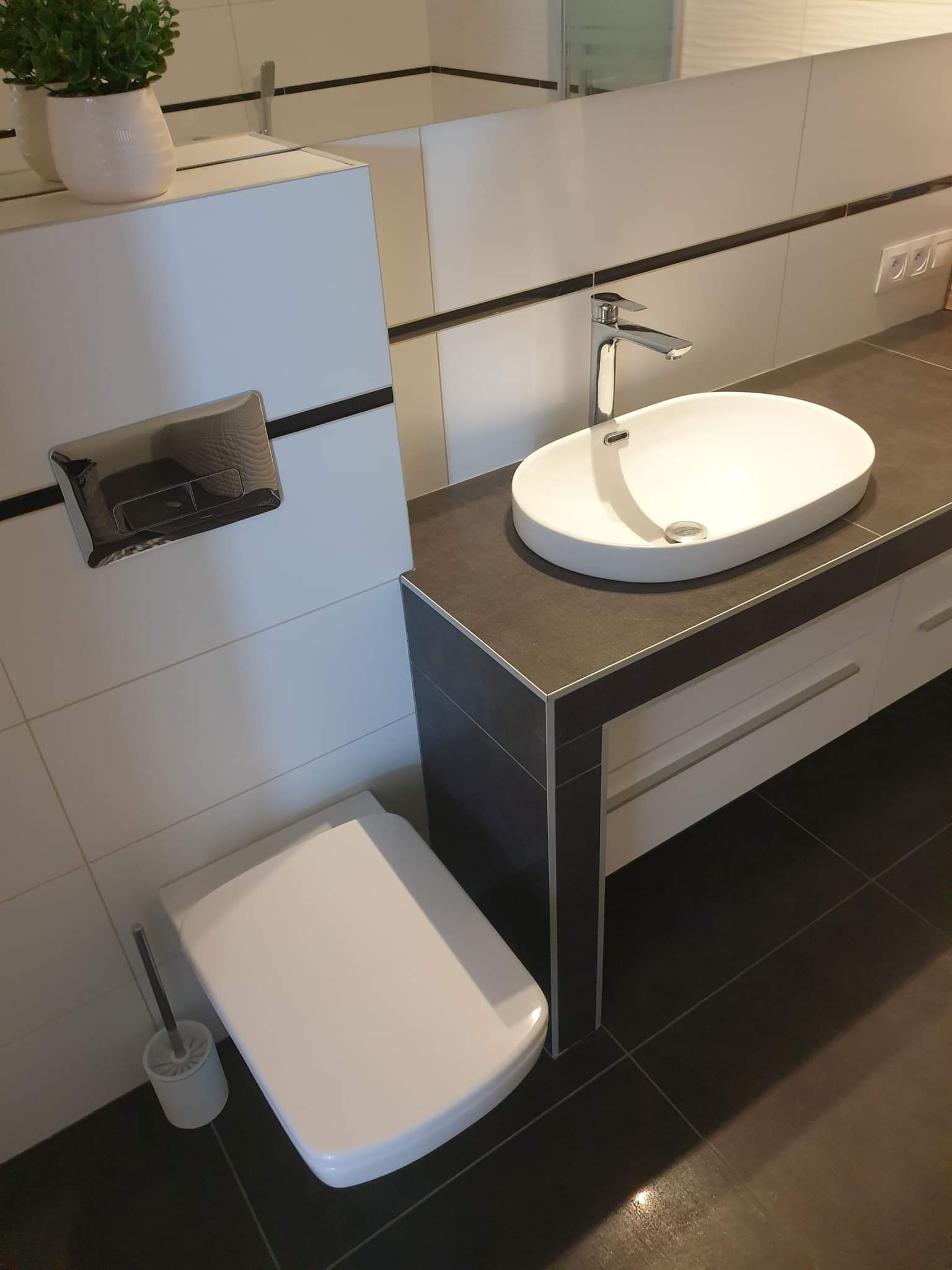 Nowoczesna łazienka z wiszącą toaletą, umywalką nablatową i ciemną, gładką podłogą. Na blacie umywalki minimalistyczna bateria. Na szafce pod umywalką białe fronty z długimi, poziomymi uchwytami.