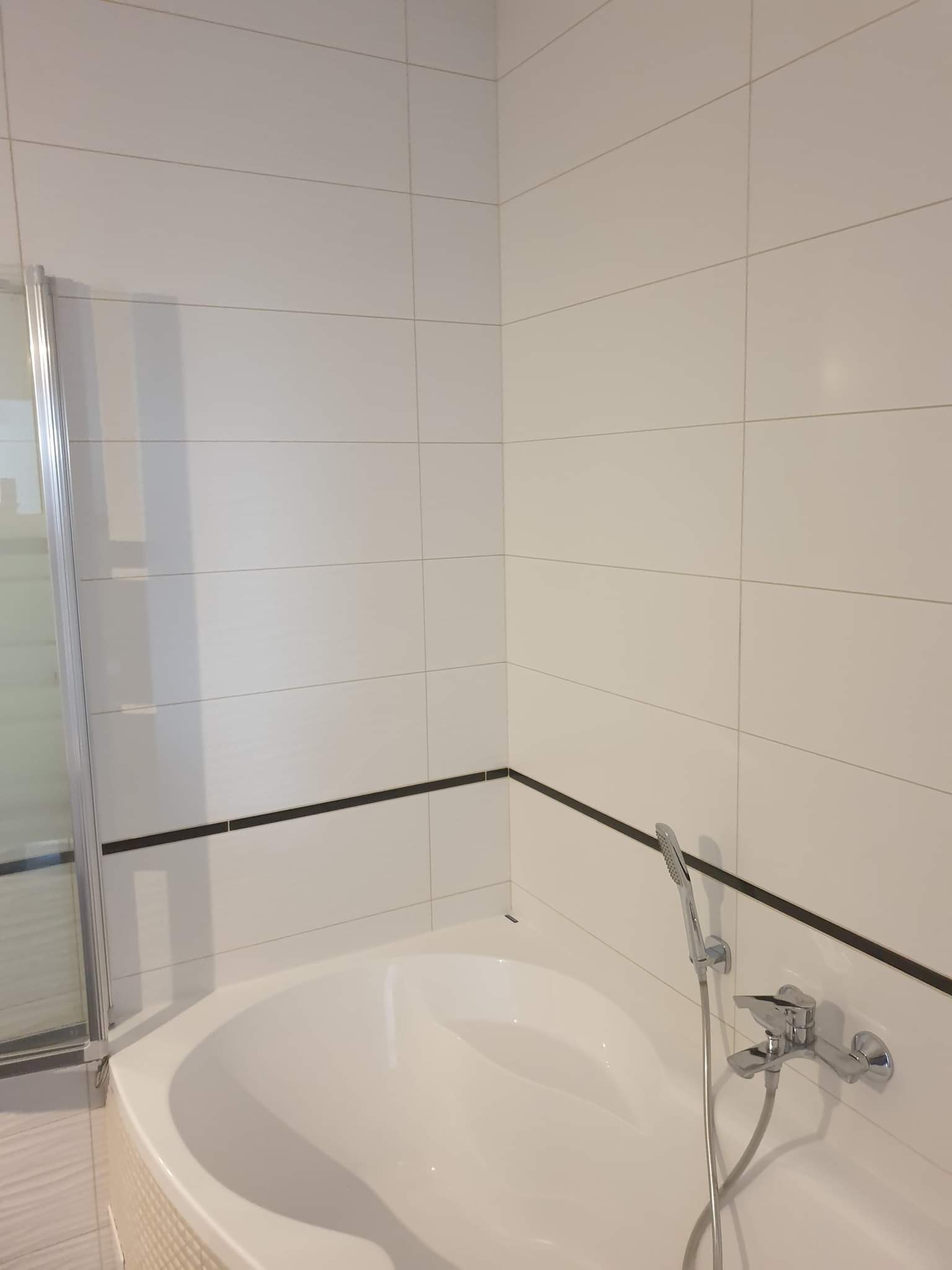 Biała, narożna wanna z chromowaną baterią i słuchawką prysznicową na tle białych płytek z czarnym paskiem. Widoczna częściowo szklana kabina prysznicowa.