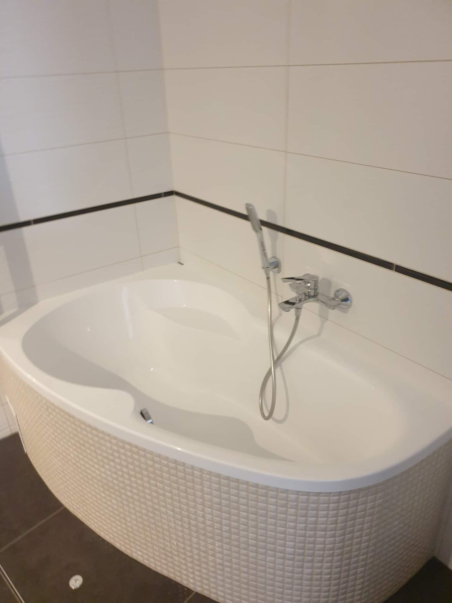 Narożna wanna obudowana mozaiką w beżowym odcieniu, z chromowaną armaturą i prysznicem na tle białych płytek z czarnym akcentem.