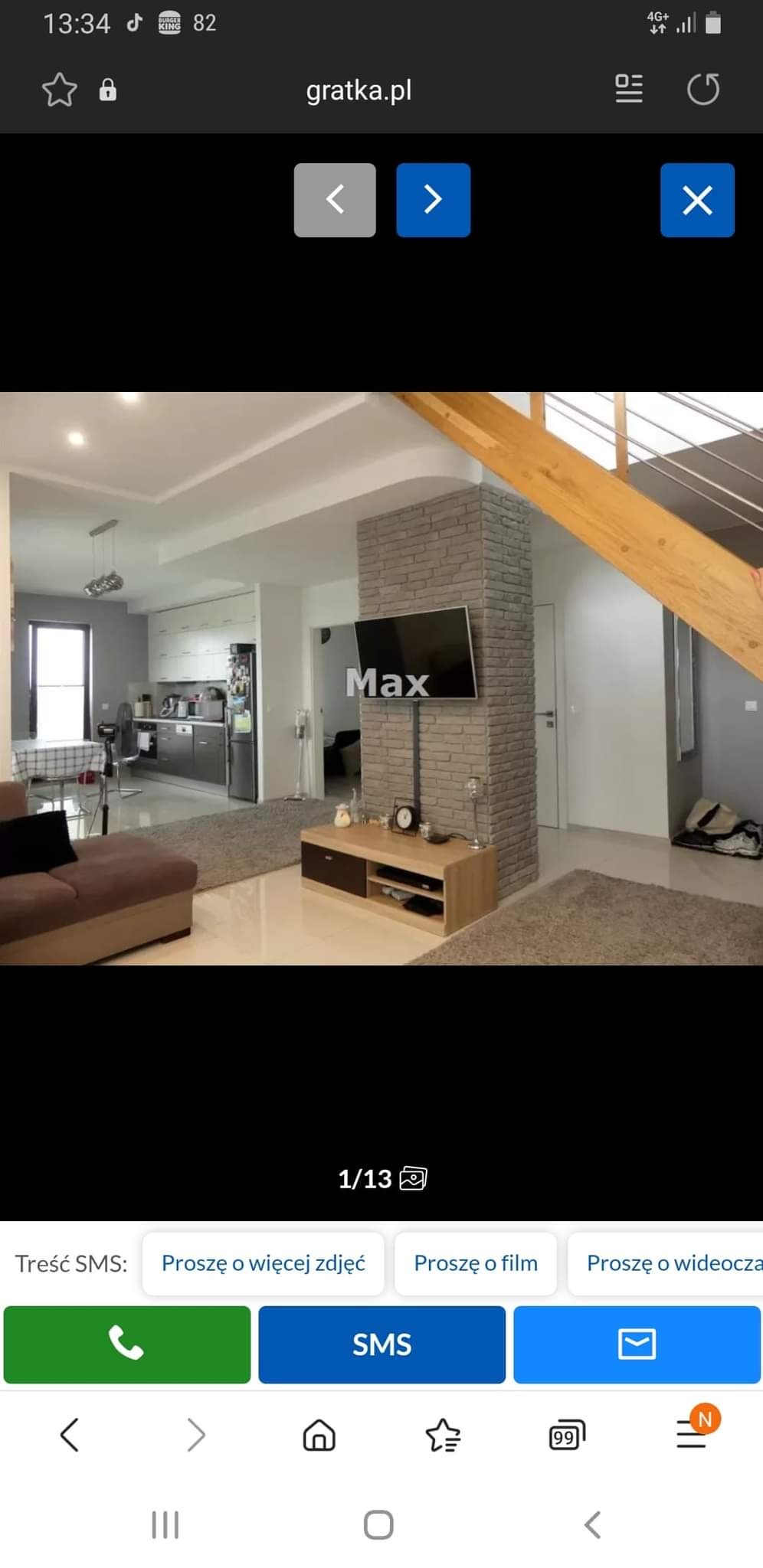 Jasne wnętrze z otwartą kuchnią i salonem, ceglany filar z telewizorem, drewniane schody z metalową balustradą, beżowe dywany i meble, nowoczesne oświetlenie.