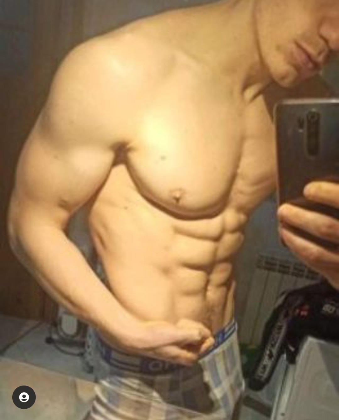 Mężczyzna robiący selfie, prezentujący wyrzeźbione ciało, widoczne mięśnie brzucha i klatki piersiowej, w tle fragment grzejnika i podłogi.