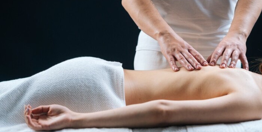 Kobieta leżąca na brzuchu pod białym ręcznikiem, podczas gdy masażysta w białej koszulce wykonuje masaż pleców. Widoczne dłonie masażysty i górna część pleców kobiety.