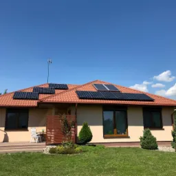 Parterowy dom jednorodzinny z panelami słonecznymi na dachu z dachówki ceramicznej, antena telewizyjna na szczycie, ogród z trawnikiem i iglakami na pierwszym planie.