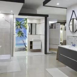 Wystawa łazienek w salonie: kabina prysznicowa, umywalka nablatowa na postumencie, szafka z umywalką w szarym kolorze i okrągłe lustro na ścianie z dekoracyjną mozaiką, płytki z motywem plaży i palm.