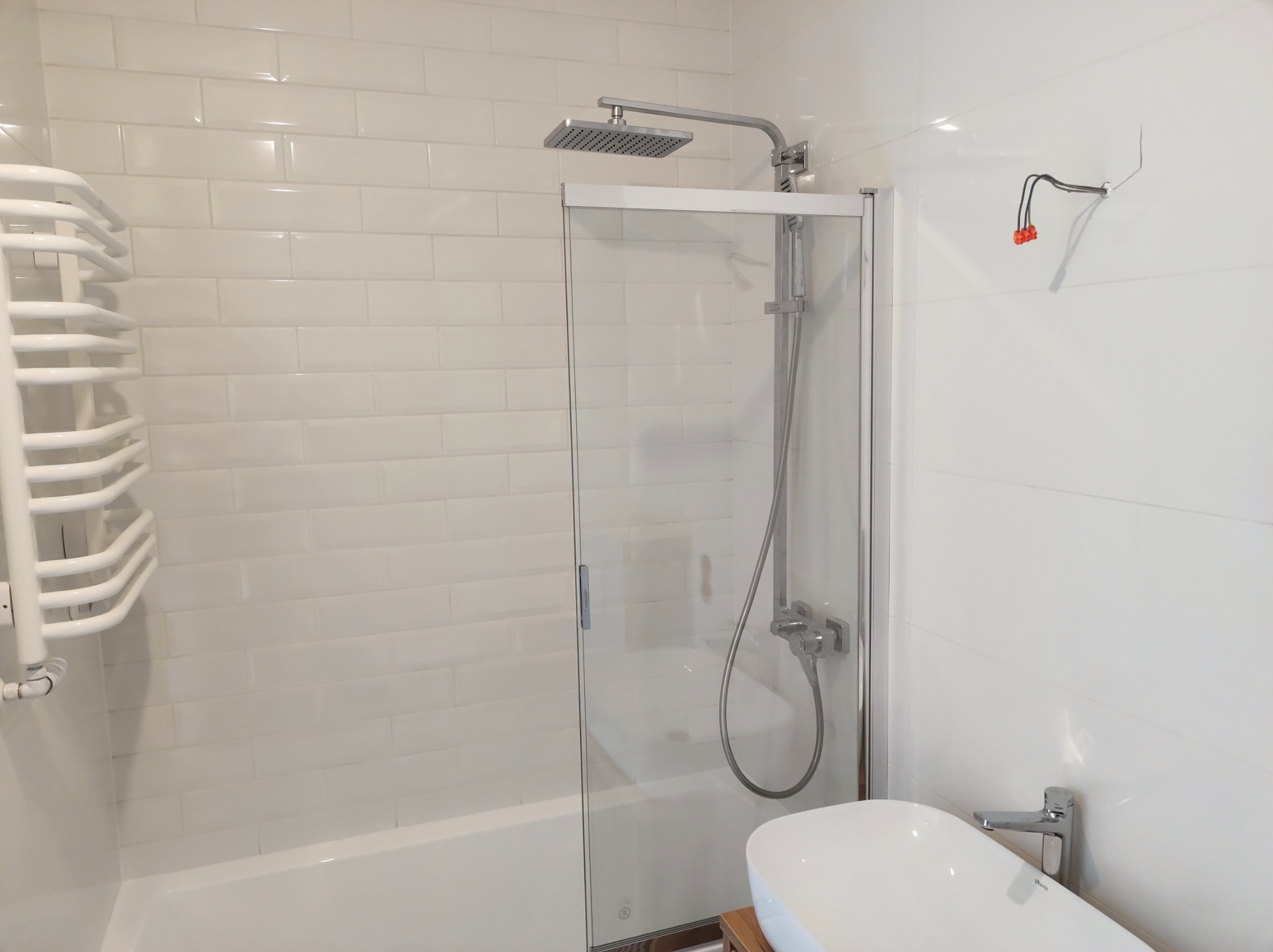 Nowoczesna, biała łazienka z kabiną prysznicową ze szklanymi drzwiami, grzejnikiem drabinkowym i niezakończonym podłączeniem elektrycznym na ścianie.