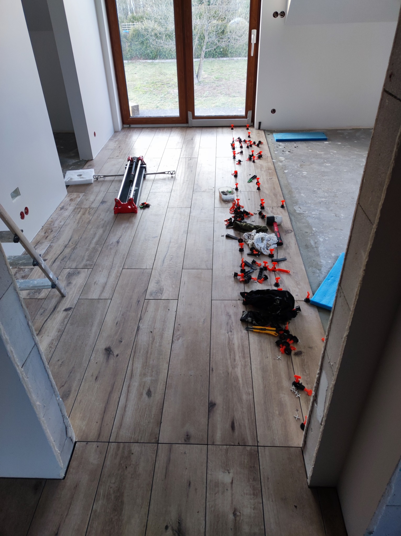 Układanie płytek imitujących drewno na podłodze w pokoju, widoczne narzędzia glazurnicze i system poziomowania płytek.