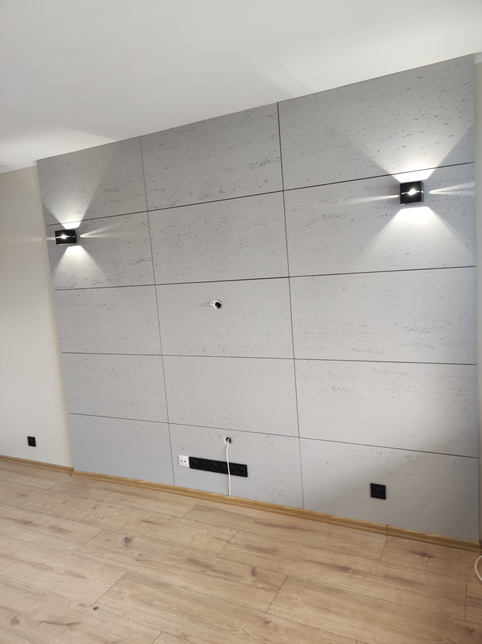 Ściana w salonie wykończona panelami imitującymi beton, podświetlona dwoma czarnymi kinkietami, z widocznymi gniazdkami elektrycznymi i otworami na kable.