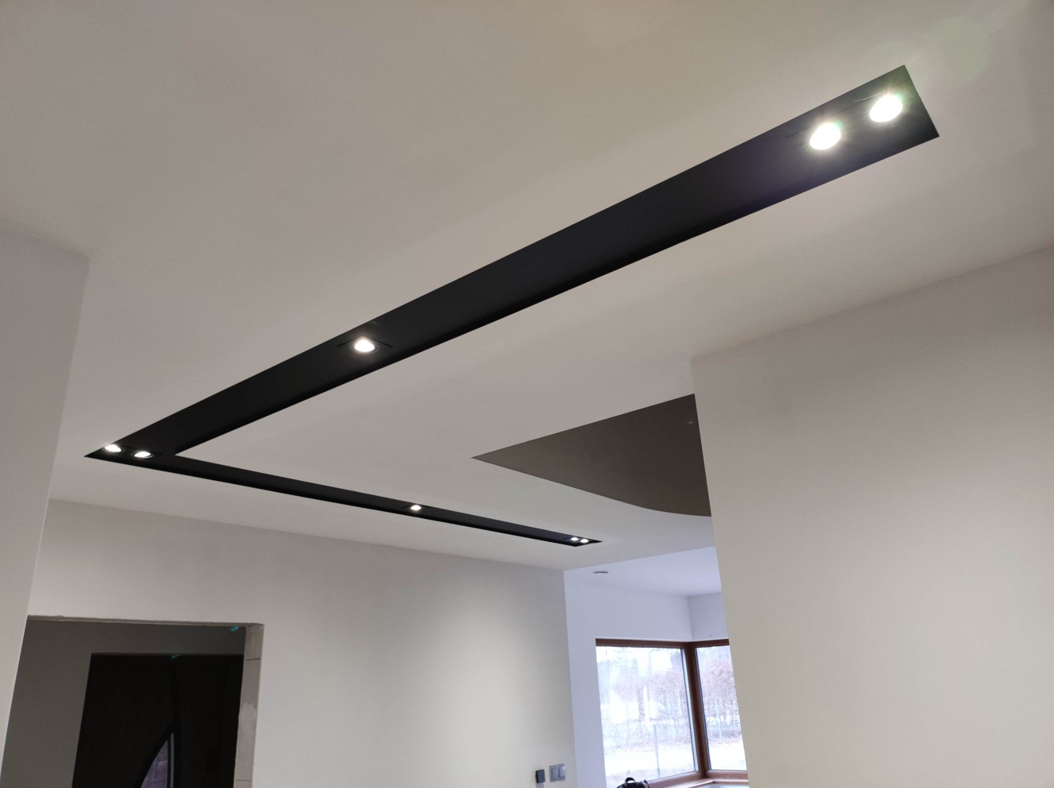 Nowoczesny sufit podwieszany z czarnymi wnękami i punktowym oświetleniem LED w jasnym wnętrzu w Solcu Kujawskim, widok z dołu.