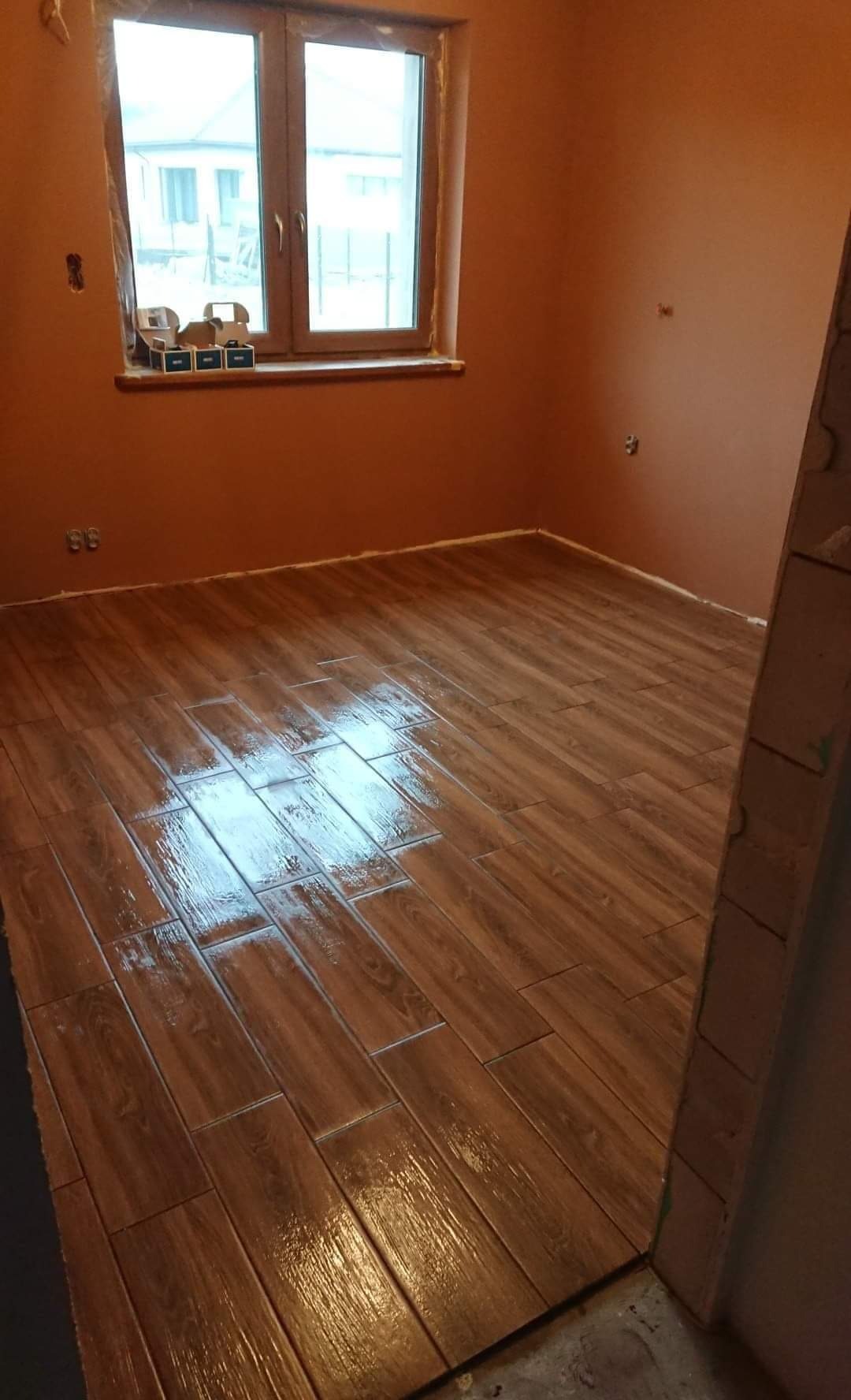 Świeżo ułożona, błyszcząca podłoga z płytek imitujących drewno w pustym pokoju z oknem, widoczne puszki po farbie na parapecie.