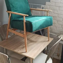 Szmaragdowy fotel z drewnianymi nóżkami i podłokietnikami, ustawiony na stole w mieszkaniu.