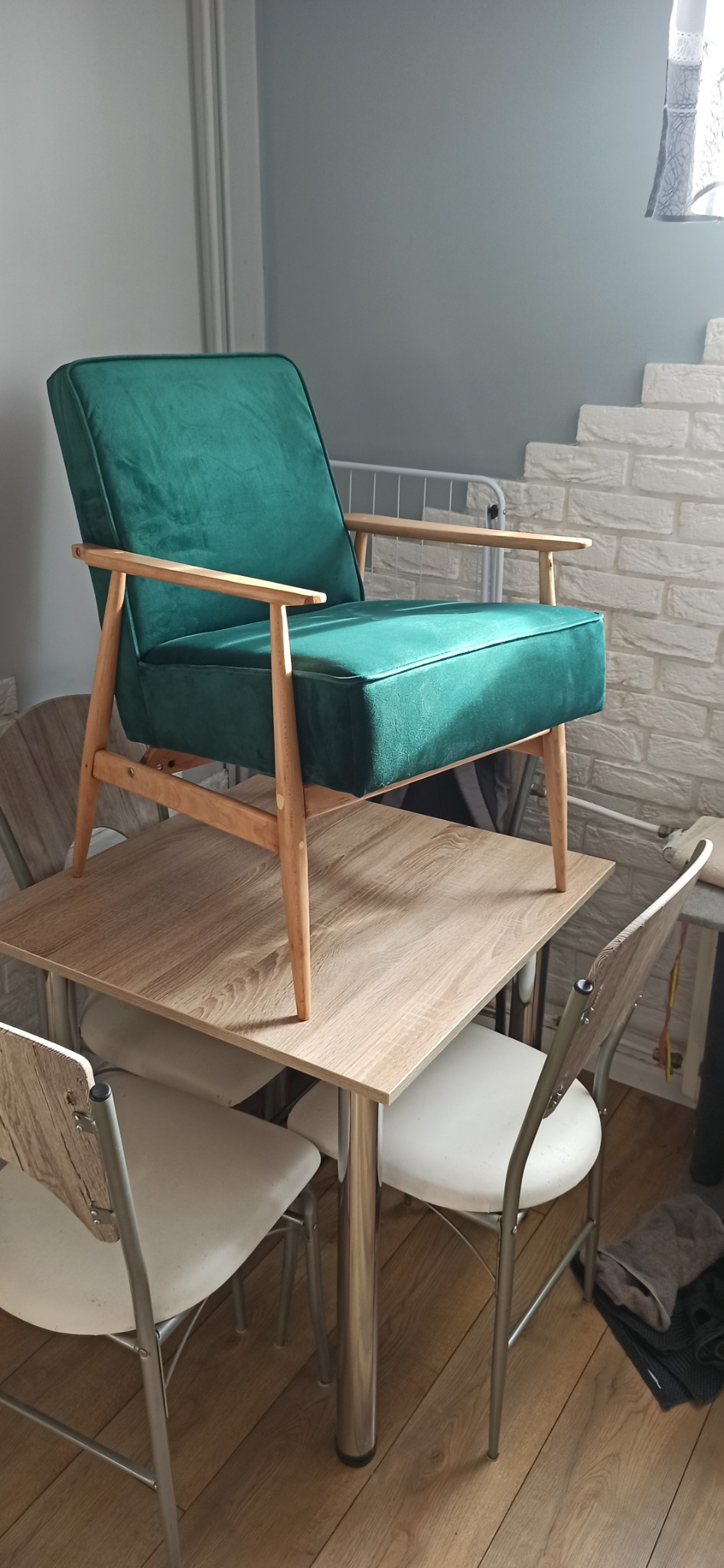 Szmaragdowy fotel z drewnianymi nóżkami i podłokietnikami, ustawiony na stole w mieszkaniu.