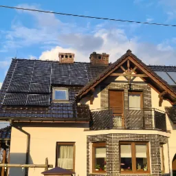 Dom jednorodzinny z zamontowanymi panelami fotowoltaicznymi na dachu z brązowej dachówki, widoczne kominy i okna dachowe, na pierwszym planie drewniana altanka z zielonym dachem, błękitne niebo...