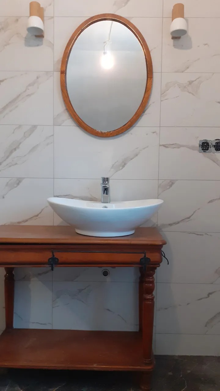 Umywalka nablatowa w kształcie łódki na drewnianej konsoli, owalne lustro w drewnianej ramie, marmurowe płytki ścienne i nowoczesne lampy ścienne.