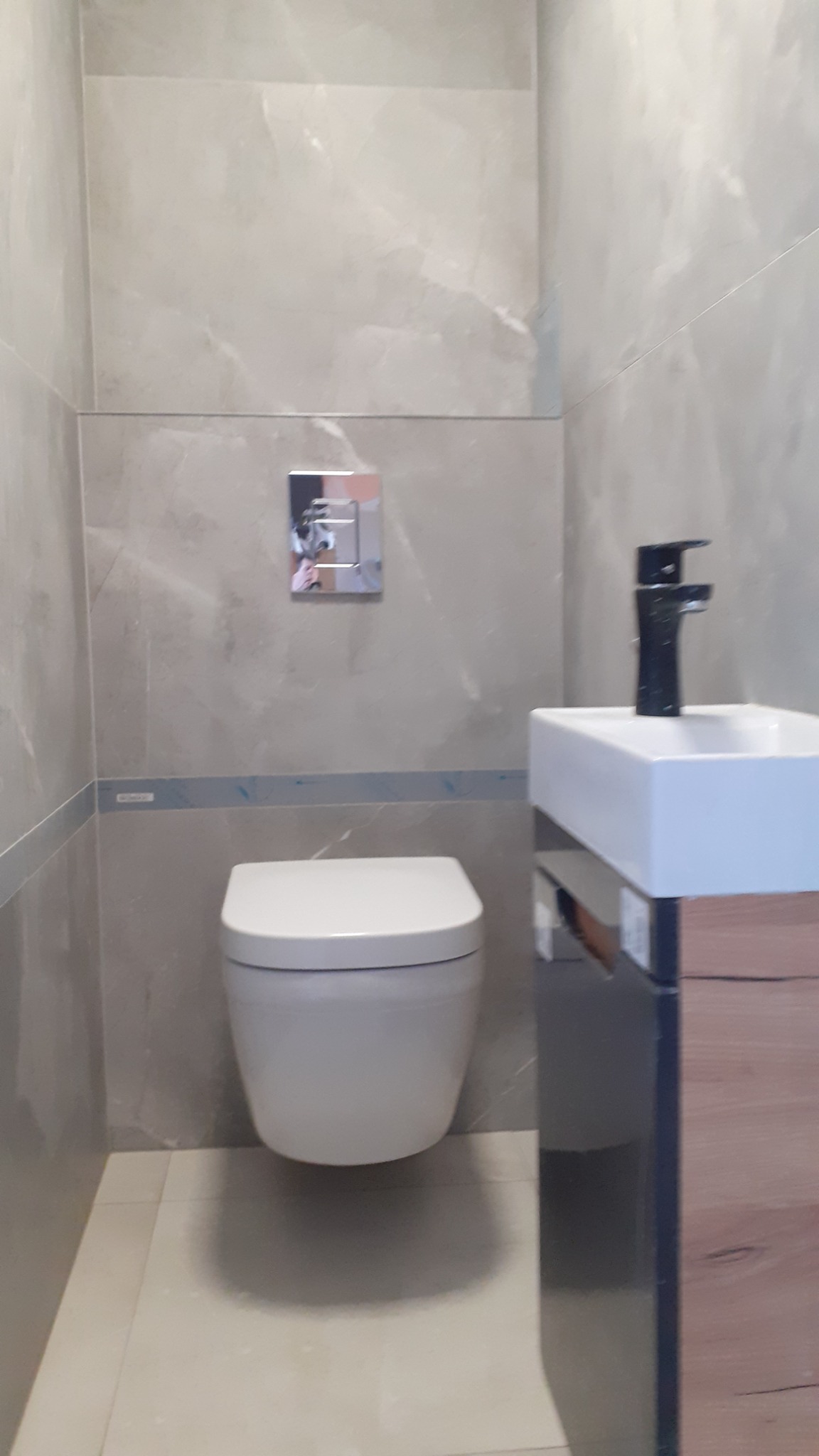 Nowoczesna łazienka z szarymi płytkami imitującymi marmur, wisząca toaleta z chromowanym przyciskiem spłukiwania i umywalka nablatowa z czarną baterią.