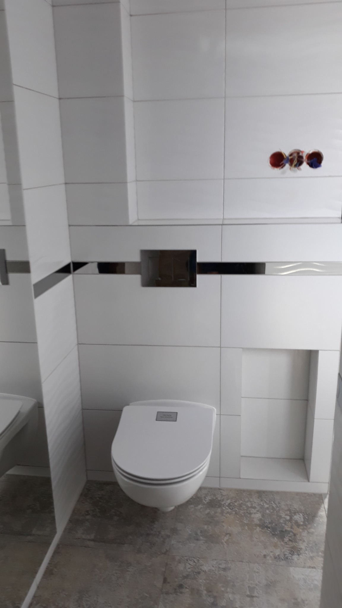 Nowoczesna toaleta wisząca w łazience wykończonej białymi płytkami z dekoracyjnym pasem i wnęką, na podłodze płytki imitujące kamień.